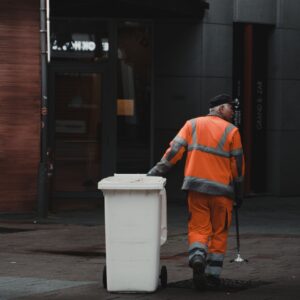 man in orange jacket and black pants standing beside white plastic trash bin